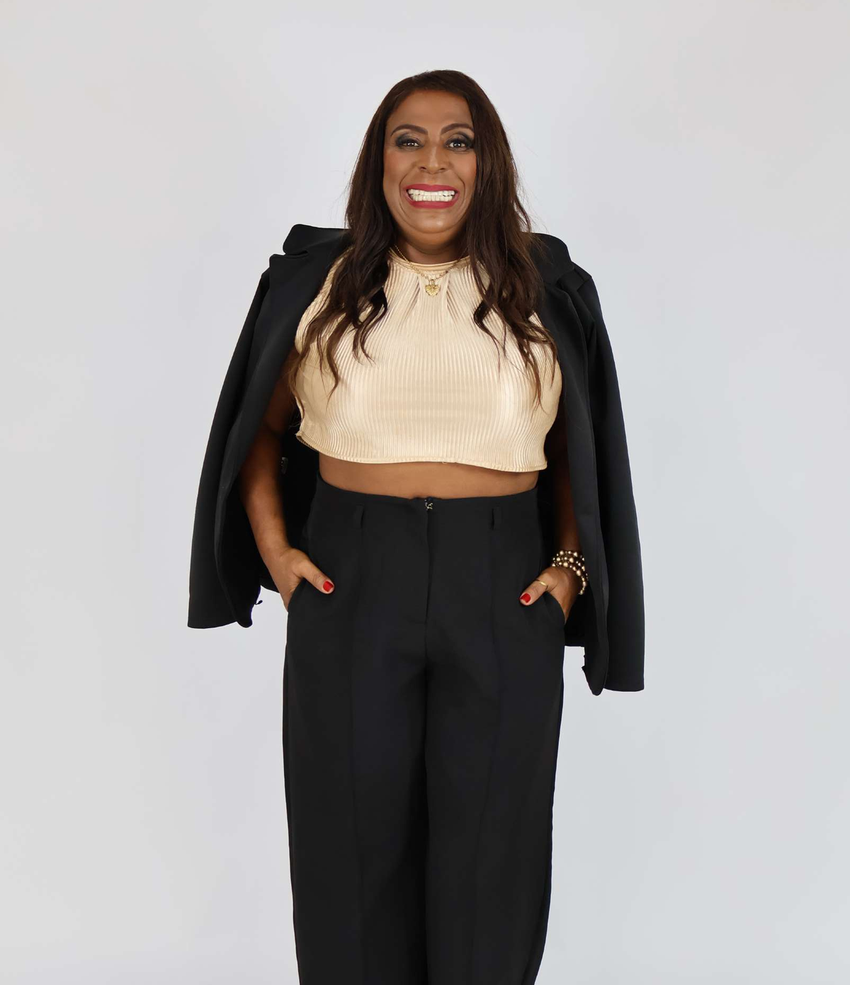 Kenia de corpo inteiro, sorrindo, com blazer preto sobre os ombros e calça preta.